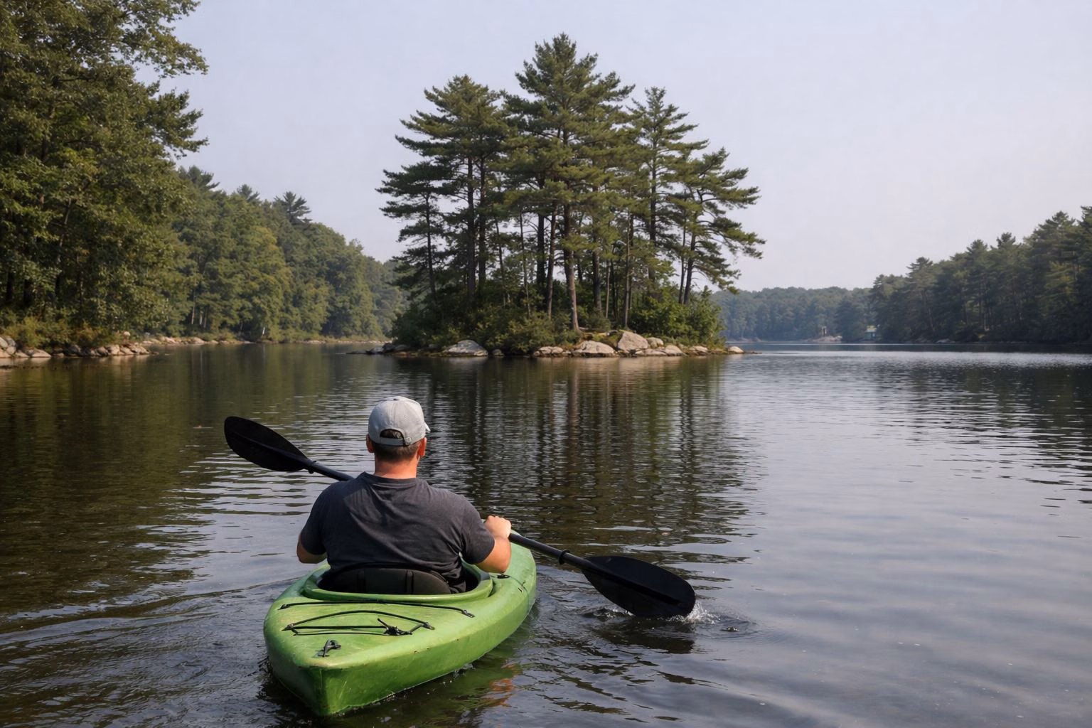 A kayaker seen from behind paddling a calm wooded lake toward a small pine island